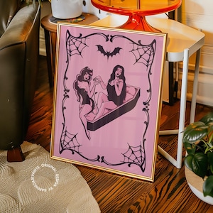 Puede incluir: Una impresión artística enmarcada con un borde dorado. La obra de arte presenta a dos mujeres en un ataúd, con un fondo rosa y telarañas negras y un diseño de murciélago. La impresión se exhibe en un suelo de madera.
