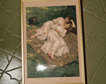 May include: Framed print featuring two women in white dresses reclining on a blanket in a grassy area. The artwork has a soft, painterly style with a gold-colored frame. The scene evokes a sense of leisure and tranquility.
