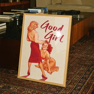 May include: A framed print featuring a vintage-style illustration of two women. One woman is standing, and the other is kneeling. The words "Good Girl" are written in red script. The frame is made of light-colored wood.