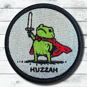 May include: Embroidered patch featuring a green frog wearing a red cape and holding a sword. The patch has a black border and the word "Huzzah" at the bottom. The frog is the central focus of the design.