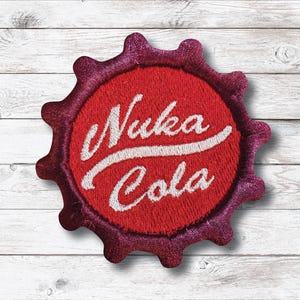 May include: Embroidered patch featuring the Nuka Cola logo. The patch is shaped like a bottle cap, with a red center and a dark pink border. The words "Nuka Cola" are written in white script across the center.