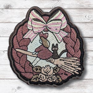 Könnte beinhalten: Gestickter Aufnäher mit einer Hexe auf einem Besen mit einer schwarzen Katze, umgeben von einem Kranz und einer großen Schleife. Der Aufnäher ist in Braun-, Rosa- und Cremetönen gehalten, mit detailliertem Design und dekorativem Rand.