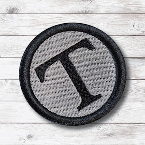 Puede incluir: Un parche redondo bordado con la letra "T" negra sobre un fondo gris. La letra está centrada dentro de un círculo, rodeada por un borde negro. El parche es para ropa o accesorios.