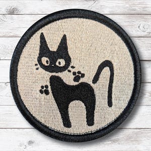 May include: Round embroidered patch featuring a black cat silhouette with large eyes and paw prints. The cat is set against a beige background, framed by a black border. The patch is likely for clothing or accessories.