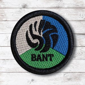 May include: Round embroidered patch with a black border. The design features a black abstract shape over a segmented background of white, blue, and green. The word "BANT" is printed in black on the green section.