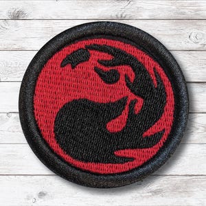 May include: Round embroidered patch featuring a red and black graphic design. The patch has a black border and a textured appearance. The central design is a stylized, flame-like shape.