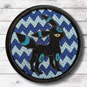 Puede incluir: Parche bordado circular con un diseño de criatura negra, ojos amarillos y detalles azules, sobre un patrón de zigzag azul y azul claro. El parche tiene un borde negro. El diseño está probablemente inspirado en una serie animada popular.