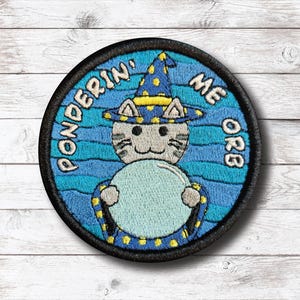 Könnte beinhalten: Runder, gestickter Aufnäher mit einer Katze, die einen Zaubererhut trägt und eine Kristallkugel hält. Der Aufnäher hat einen schwarzen Rand und den Text "Ponderin' Me Orb". Die Katze ist grau, der Hut blau mit gelben Punkten.