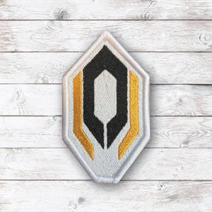 May include: A diamond-shaped embroidered patch featuring a central black hexagon outline surrounded by two vertical gold bars on a light gray background, all enclosed within a thick white border with a textured, stitched fabric finish.