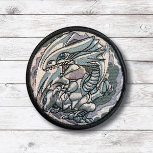 Puede incluir: Parche bordado circular que representa un dragón azul y blanco con un borde negro. El dragón es el foco principal, con detalles intrincados en su diseño. El fondo presenta tonos de azul, gris y morado, agregando profundidad al parche.