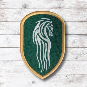 Può includere: Una toppa ricamata a forma di scudo con uno sfondo verde scuro e un design stilizzato di testa di cavallo argentata con criniera fluente. La toppa è bordata da uno spesso filo dorato metallizzato, che crea un contorno netto per l'emblema.