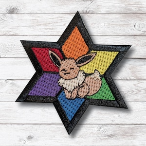 May include: Embroidered patch featuring a cartoon Eevee character inside a black star. The star is divided into colorful diamond shapes: red, orange, yellow, green, blue, and purple. The Eevee is light brown with a cream-colored chest and a smiling expression.