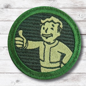 May include: A round, green embroidered patch featuring a cartoon character giving a thumbs-up. The character is in a yellow-green color, with a matching outline. The patch has a dark green background and a green border.