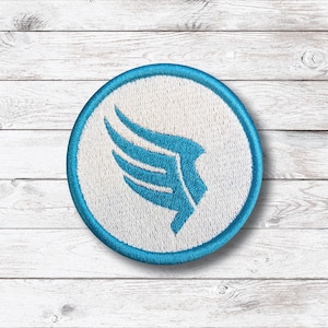 Puede incluir: Parche bordado circular con un diseño de ala azul sobre fondo blanco, enmarcado por un borde azul. El parche tiene una superficie texturizada y probablemente está hecho de tela. El fondo es un patrón de vetas de madera clara.