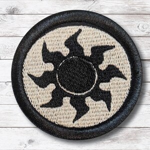 Op de afbeelding: Geborduurde ronde patch met een zwart zonnemotief op een beige achtergrond, omlijst door een zwarte rand. De zon heeft een centrale zwarte cirkel en gestileerde stralen. Voor kleding of accessoires.