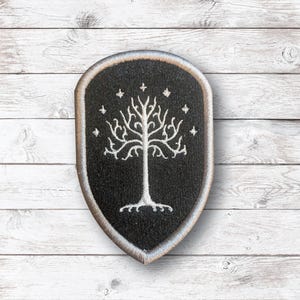 May include: A black and silver embroidered patch featuring a white tree design with stars on a shield shape. The patch has a silver border and is likely an emblem or logo.