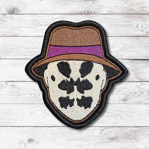 May include: Embroidered patch featuring the iconic Rorschach mask from the Watchmen comic series. The patch has a brown fedora with a purple band, a black outline, and a cream-colored face with inkblot-like patterns.