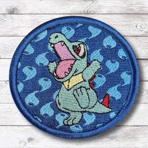May include: Embroidered patch featuring a cartoon alligator with an open mouth, showcasing pink and red details. The alligator is light green with a yellow collar and red accents. The patch has a blue border and a blue patterned background.