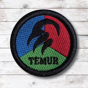 May include: Embroidered circular patch with a black border. The patch features a segmented design with blue, red, and green sections. A black claw-like graphic is centered above the word "TEMUR" in green.