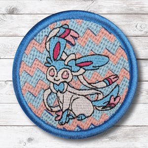 May include: Round embroidered patch featuring a cartoon character in shades of blue, pink, and white. The character is set against a light blue and pink zigzag pattern, with a blue border. The patch is likely for clothing or accessories.