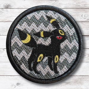 Puede incluir: Parche bordado circular que representa un personaje Umbreon negro con detalles amarillos y una boca roja. El diseño está sobre un patrón de zigzag gris y blanco, encerrado por un borde negro. El parche es de tela.