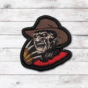 Peut inclure: Un écusson brodé représentant un personnage au visage cicatrisé portant un chapeau fedora marron et un pull rouge. Le personnage a une main griffue avec des lames métalliques étendues, le tout entouré d'une bordure noire épaisse.