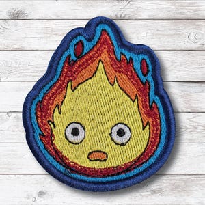 Könnte beinhalten: Gestickter Aufnäher mit einem Cartoon-Feuerdämon mit gelbem Gesicht, großen schwarzen Augen und einem orangefarbenen Mund. Die Flammen sind rot und orange, blau umrandet. Der Aufnäher befindet sich auf einer weißen Holzoberfläche.