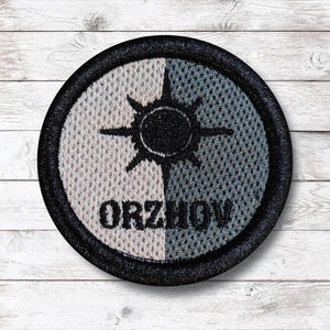 Op de afbeelding: Een ronde patch met een zwarte rand, verticaal verdeeld in twee secties. De ene kant is lichtgrijs, de andere blauwgrijs. Een zwart zonnestraalontwerp staat boven het woord "ORZHOV" in zwarte tekst.