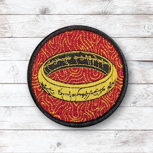 Puede incluir: Parche bordado redondo que muestra un anillo dorado con letras negras sobre un fondo rojo, con un borde negro. El anillo, con su inscripción detallada, es el punto focal, sobre un patrón texturizado.