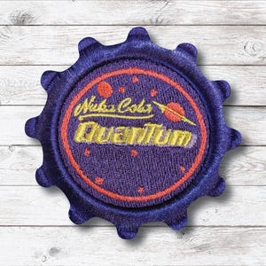 May include: Embroidered patch featuring a purple bottle cap design with a circular center. The center has the text "Nuka Cola Quantum" in yellow, with a red ring and space-themed graphics.