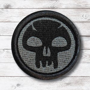 May include: Round black patch with a skull design. The skull is black with gray detailing and is centered within a gray circle. The patch has a black border and is set against a white wooden background.