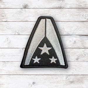 May include: A triangular embroidered patch featuring a black background with three white stars arranged in a triangle. The patch is bordered by a thick black edge and includes two light gray curved side panels that meet at the top point.