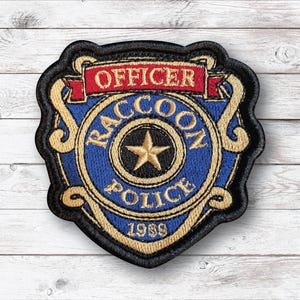 May include: Embroidered patch featuring a shield design with the words "Officer" and "Raccoon Police" in gold lettering on a blue background. A gold star is in the center, with the year "1989" at the bottom. The patch has a black border.