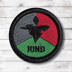 Puede incluir: Un parche redondo con borde negro, con un diseño segmentado en gris, rojo y verde. Un emblema negro está centrado y la palabra "JUND" está impresa en negro en el segmento verde.
