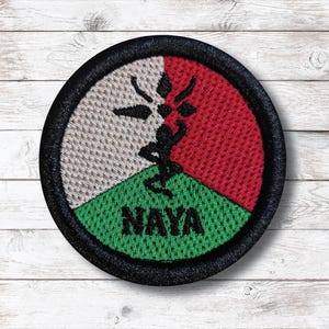 Op de afbeelding: Ronde geborduurde patch met een zwarte rand. Het ontwerp is verdeeld in drie secties: wit, rood en groen. Een zwart silhouet van een figuur en het woord "NAYA" zijn op de patch geborduurd.