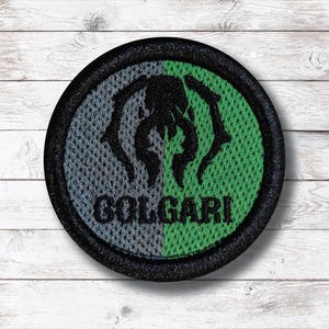 Può includere: Toppa rotonda ricamata con bordo nero. La toppa presenta un design a rete grigio e verde con una grafica nera e la scritta "COLGARI" in nero. La toppa è su una superficie di legno bianco.