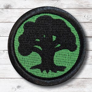 May include: Round black and green embroidered patch featuring a stylized black tree design. The tree is set against a green background within a black border. The patch is likely for clothing or accessories.