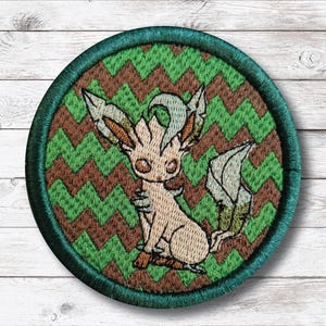 May include: Embroidered patch featuring a cartoon creature with large eyes, sitting on a green and brown zigzag pattern. The patch has a dark green border. The creature is primarily beige with green and brown accents.