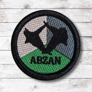 Op de afbeelding: Een ronde patch met een zwarte rand. Het toont een gestileerd zwart silhouet van twee figuren in een dynamische pose. De patch is verdeeld in drie secties: grijs, blauwgrijs en groen, met het woord "ABZAN" in groen.