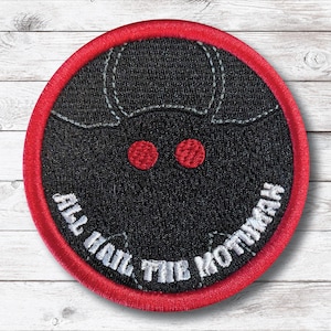 May include: Round black patch with a red border featuring a stylized Mothman design. The Mothman has red eyes and white stitched details. The text "ALL HAIL THE MOTHMAN" is in white, arched around the bottom of the patch.