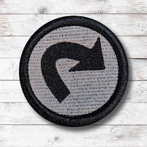 May include: Round embroidered patch featuring a black curved arrow pointing right within a white circle. The arrow is on a textured gray background, surrounded by a black border. The patch is designed for application on clothing or accessories.