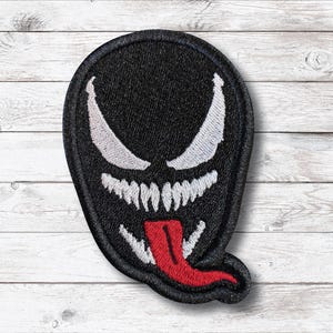 May include: Black embroidered patch featuring the face of a comic book character. The design includes white eyes and teeth, and a red tongue. The patch is an oval shape.