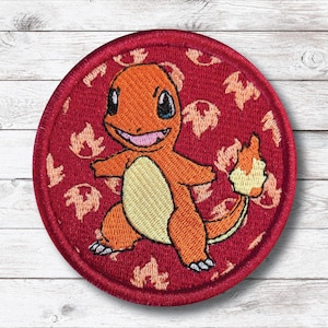 May include: A round, red embroidered patch featuring a smiling orange Charmander character with a yellow belly and a flame on its tail. The background is red with a repeating flame pattern. The patch is likely for clothing or accessories.
