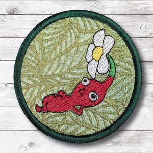 May include: Embroidered patch featuring a red creature with large eyes, holding a white flower with a yellow center. The creature is set against a light green background with a leaf pattern, framed by a dark green border. The patch is round.