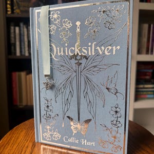 May include: A hardback book titled "Quicksilver" by Callie Hart. The book features a light blue cover with silver and gold foil detailing, including butterflies, flowers, and a sword design. A light blue ribbon bookmark with a butterfly charm is attached.