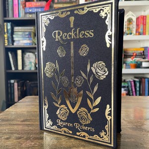 May include: A black hardcover book with gold foil lettering and illustrations. The title is "Reckless" by Lauren Roberts.