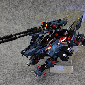 May include: A blue and black robot toy with a wolf-like head and a large gun attached to its back. The robot has red and blue lights on it.