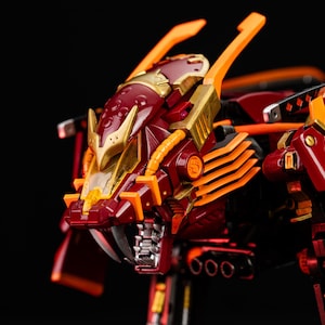 May include: A detailed close-up of a red and gold robotic figure, possibly a toy or model. The figure has a stylized design with sharp angles, orange accents, and a metallic finish. The head features a detailed face with sharp teeth.