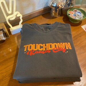 Peut inclure: Un t-shirt gris anthracite plié avec les mots "TOUCHDOWN Kansas City" en lettres orange et rouge. Le t-shirt est empilé sur d'autres t-shirts pliés. L'arrière-plan comprend une lumière en forme de signe de paix et des décorations de boule disco.