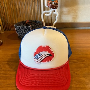 May include: A red, white, and blue trucker hat with a red brim, white front panel, and blue mesh back. The hat features an embroidered design of red lips with an American flag inside.